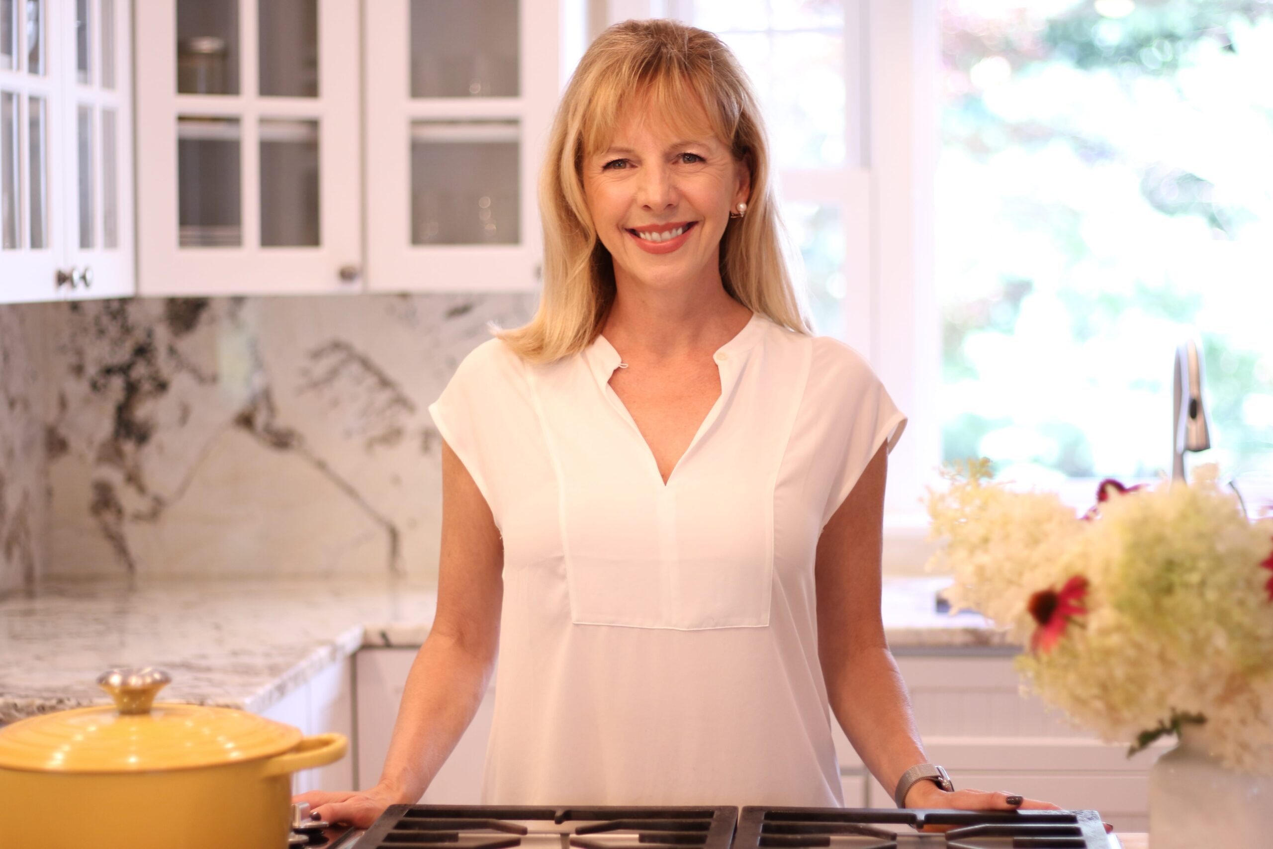 Lindsey smiling in the kitchen next to a yellow pot, wearing a white top a